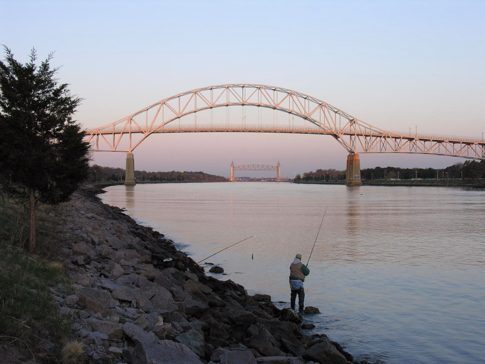 A person fishing from the rocks of the Cape Cod Canal at sunset, with the canal's bridges in the distance