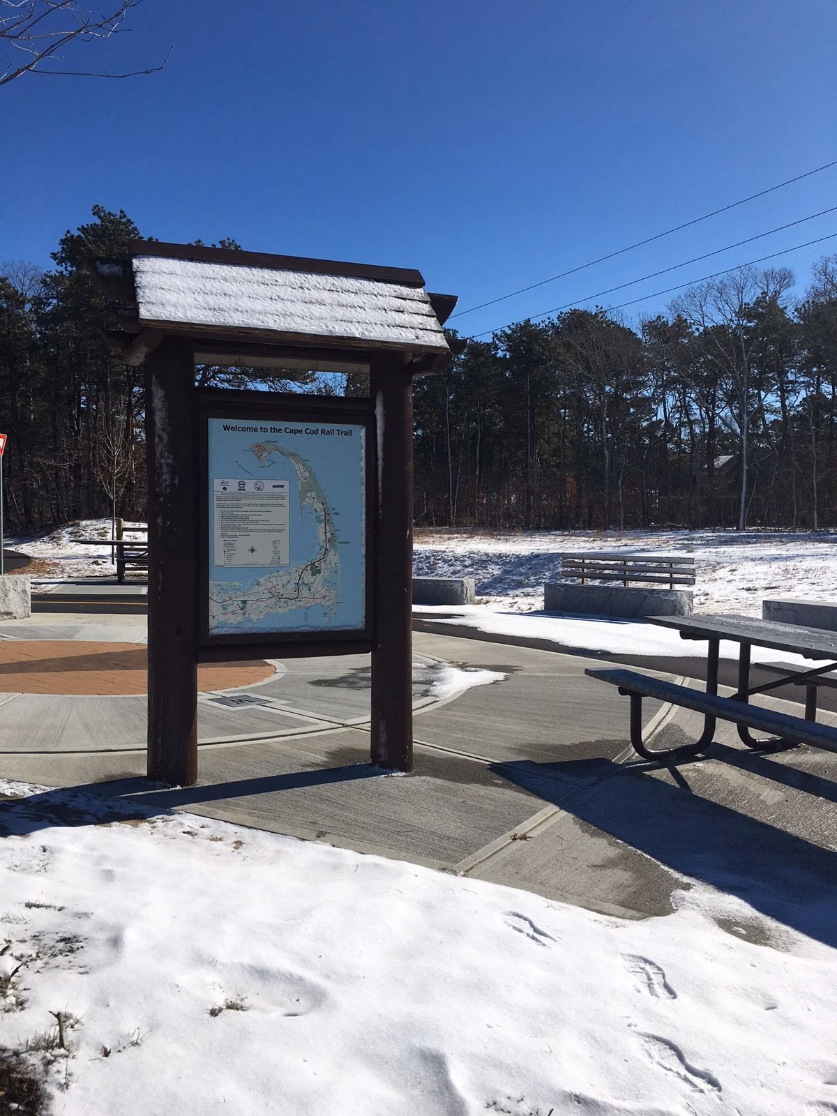 A trail map kiosk and picnic table at a Cape Cod Rail Trail trailhead in Dennis in winter